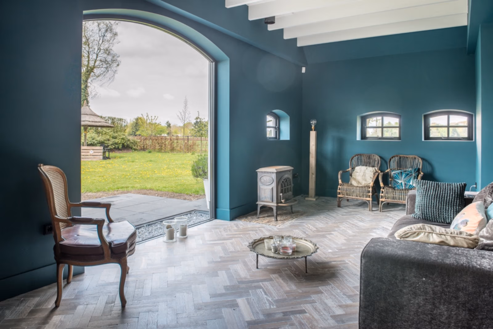 Een elegante woonkamer met diepe blauwe muren, een boogvormig raam dat uitzicht biedt op een groene tuin, en een visgraat houten vloer.