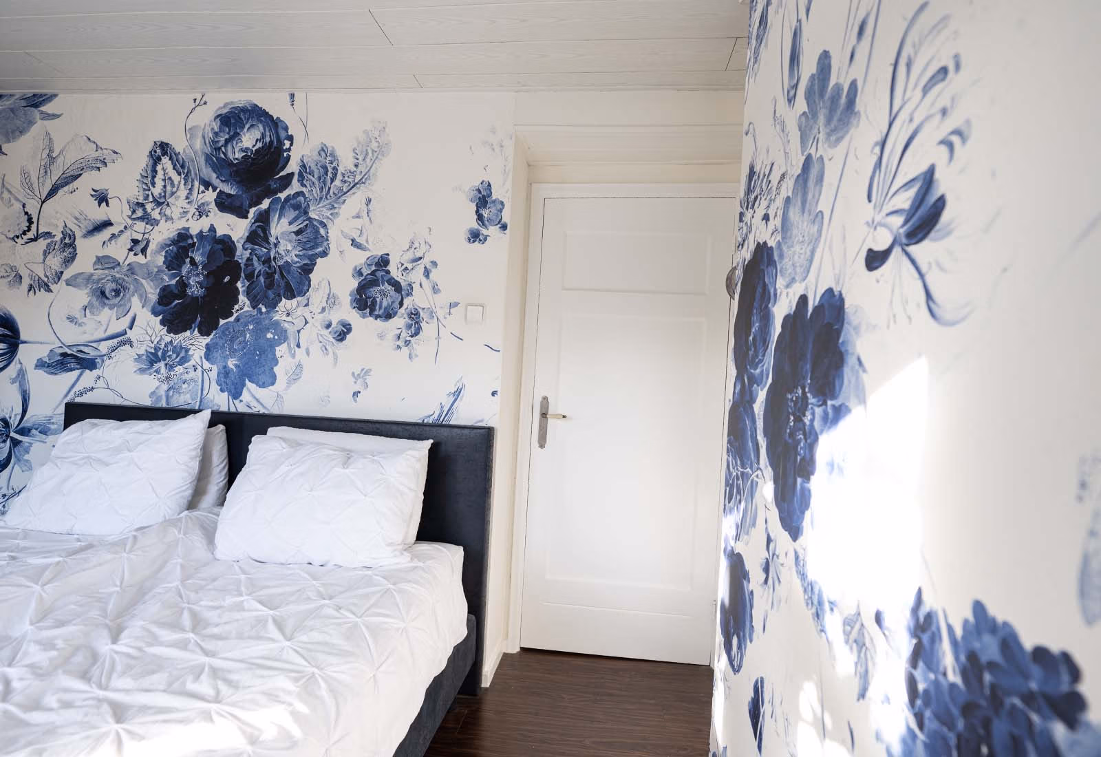 Slaapkamer met blauw-wit bloemmotiefbehang op de muren, een tweepersoonsbed met witte beddengoed, en een deur in klassieke stijl.