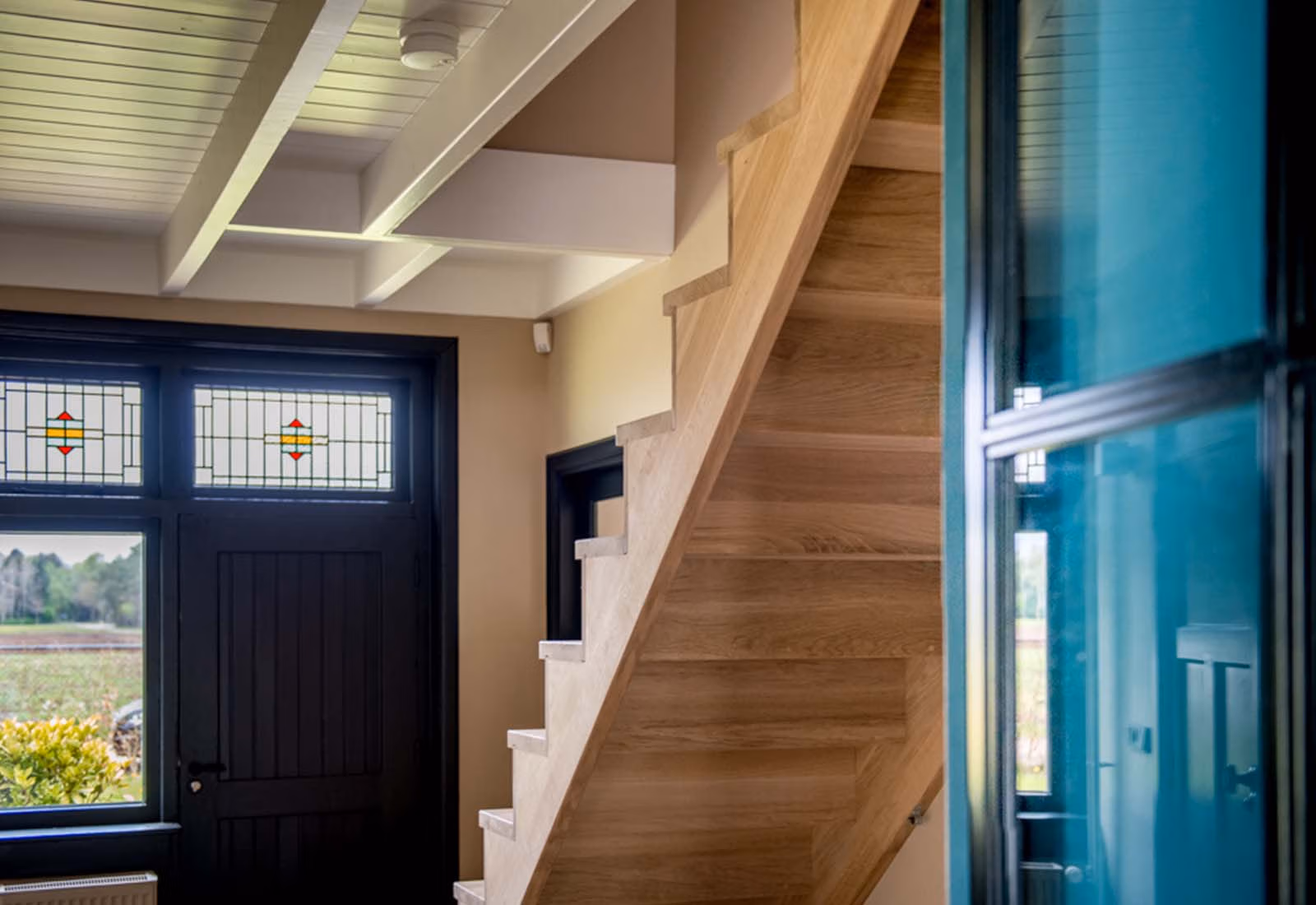Moderne hal met een houten trap, zwartgelakte deuren met glas-in-loodramen en een strak wit plafond met zichtbare balken.