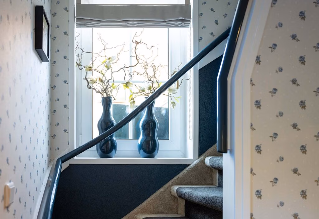Bovenaanzicht van een trap met bloemmotiefbehang op de muren, een zwartgelakte trapleuning en een raam met decoratieve takken in blauwe vazen.
